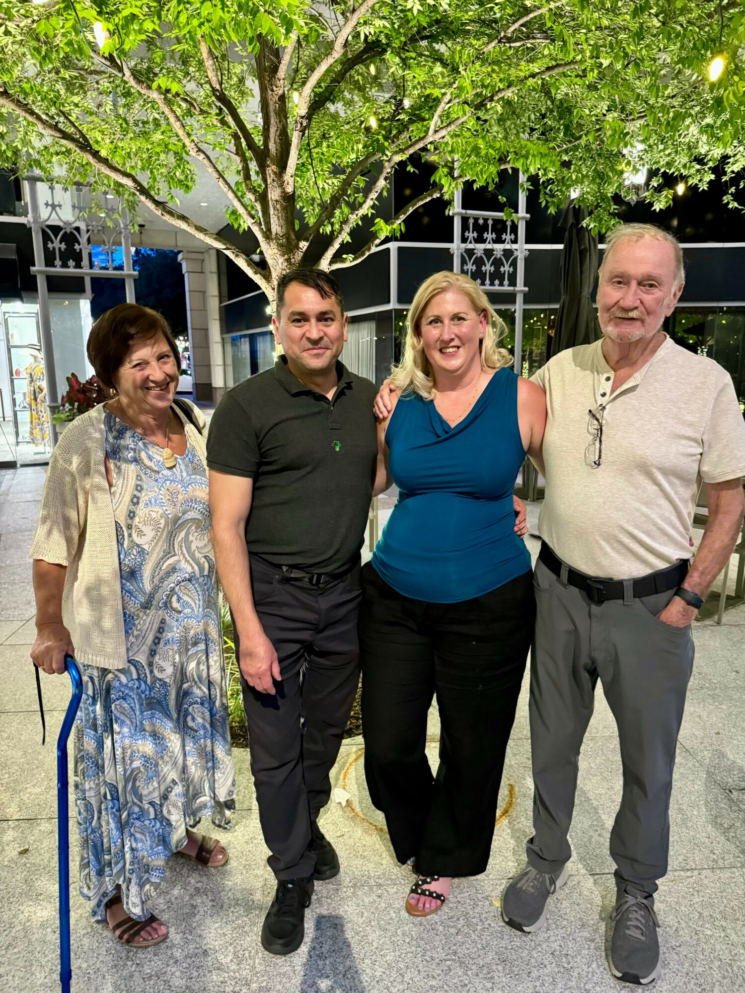 Four people standing outdoors at night, smiling, with a tree and decorative fence in the background.