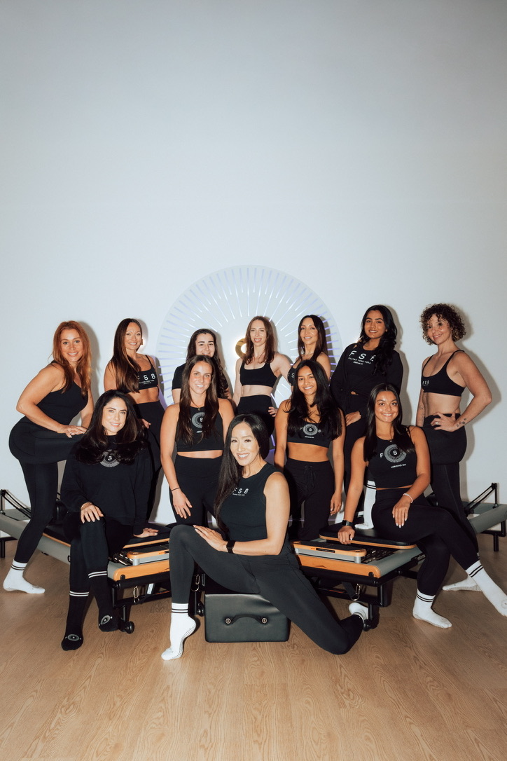Group of ten women in black outfits posing in a room with a white wall and wooden floor.