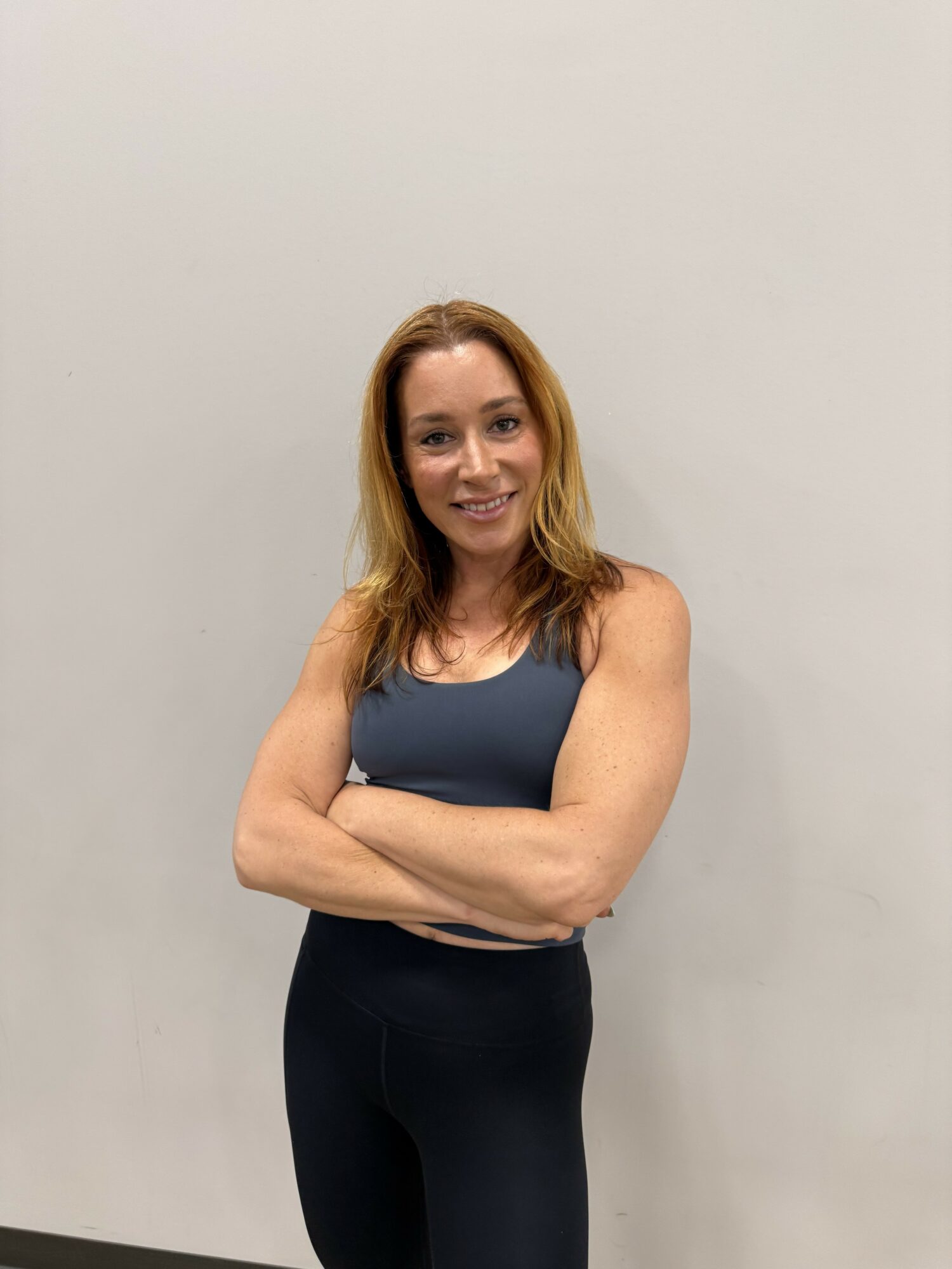 Woman with shoulder-length hair smiling, arms crossed, wearing a sleeveless top and dark pants, standing against a plain wall.