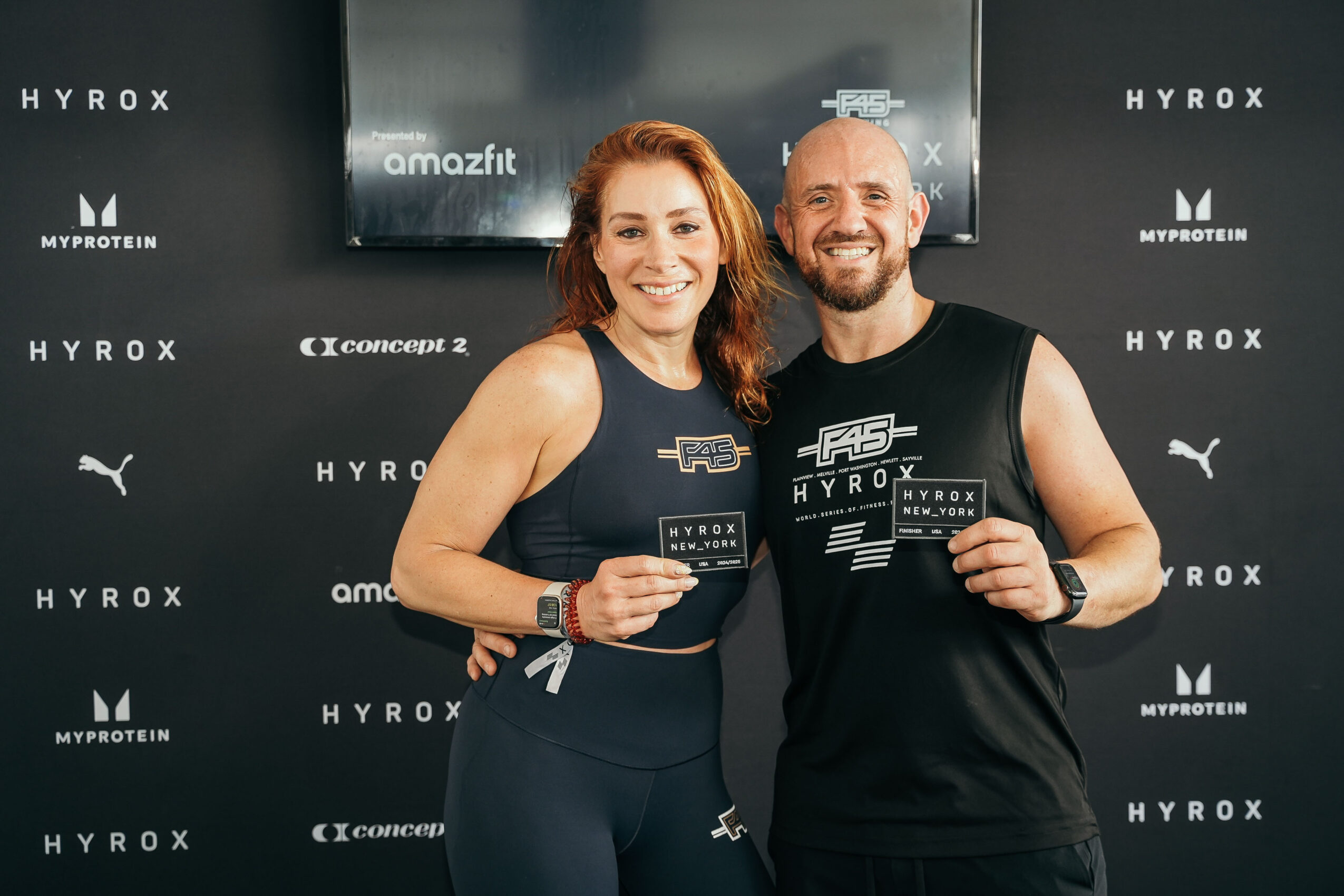Two smiling people holding certificates, standing in front of a black backdrop with fitness brand logos.