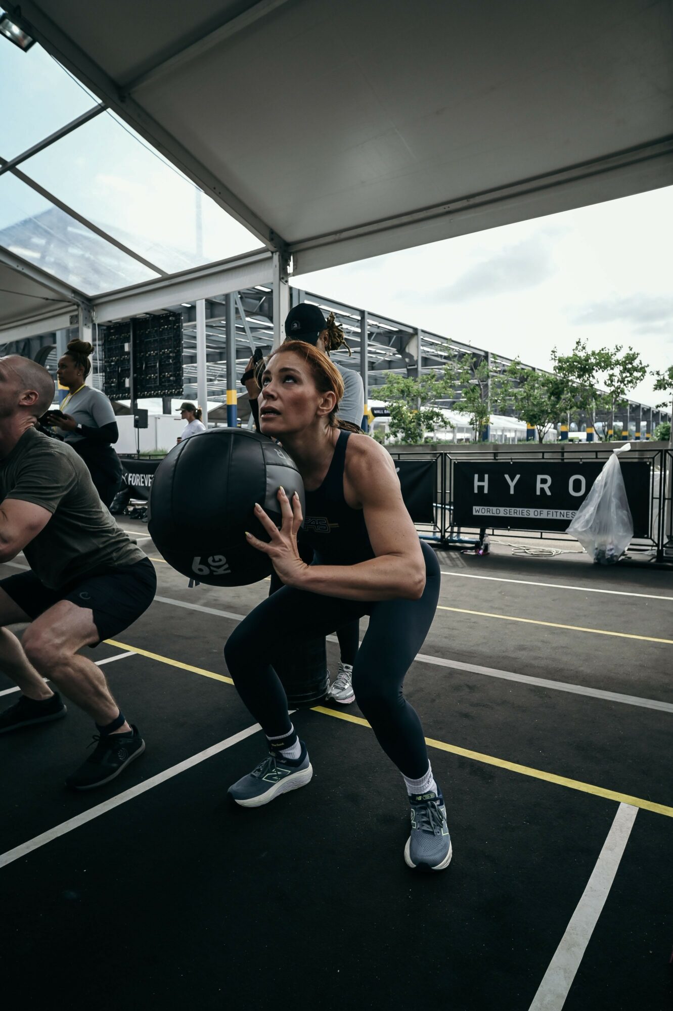 Woman holding a medicine ball in a squat position during a workout session under a tent, with people and buildings outside.