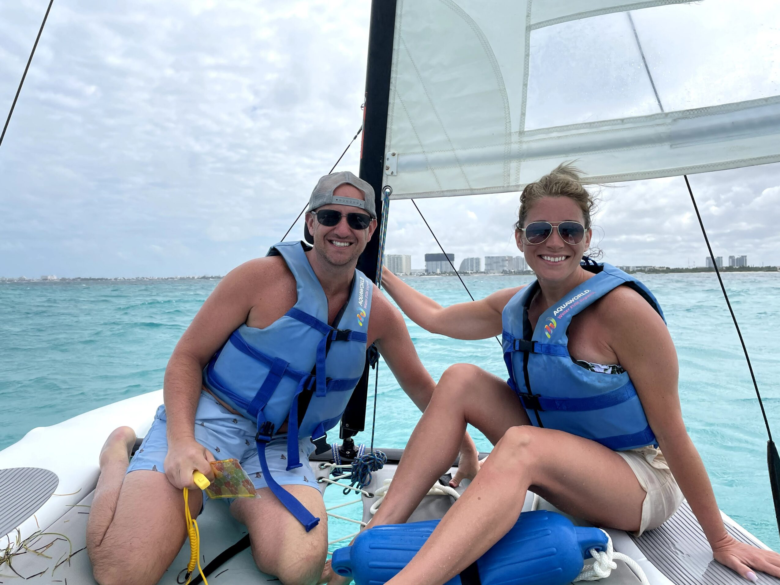 Two smiling women in life jackets sit on a sailboat, with water and sky in the background.