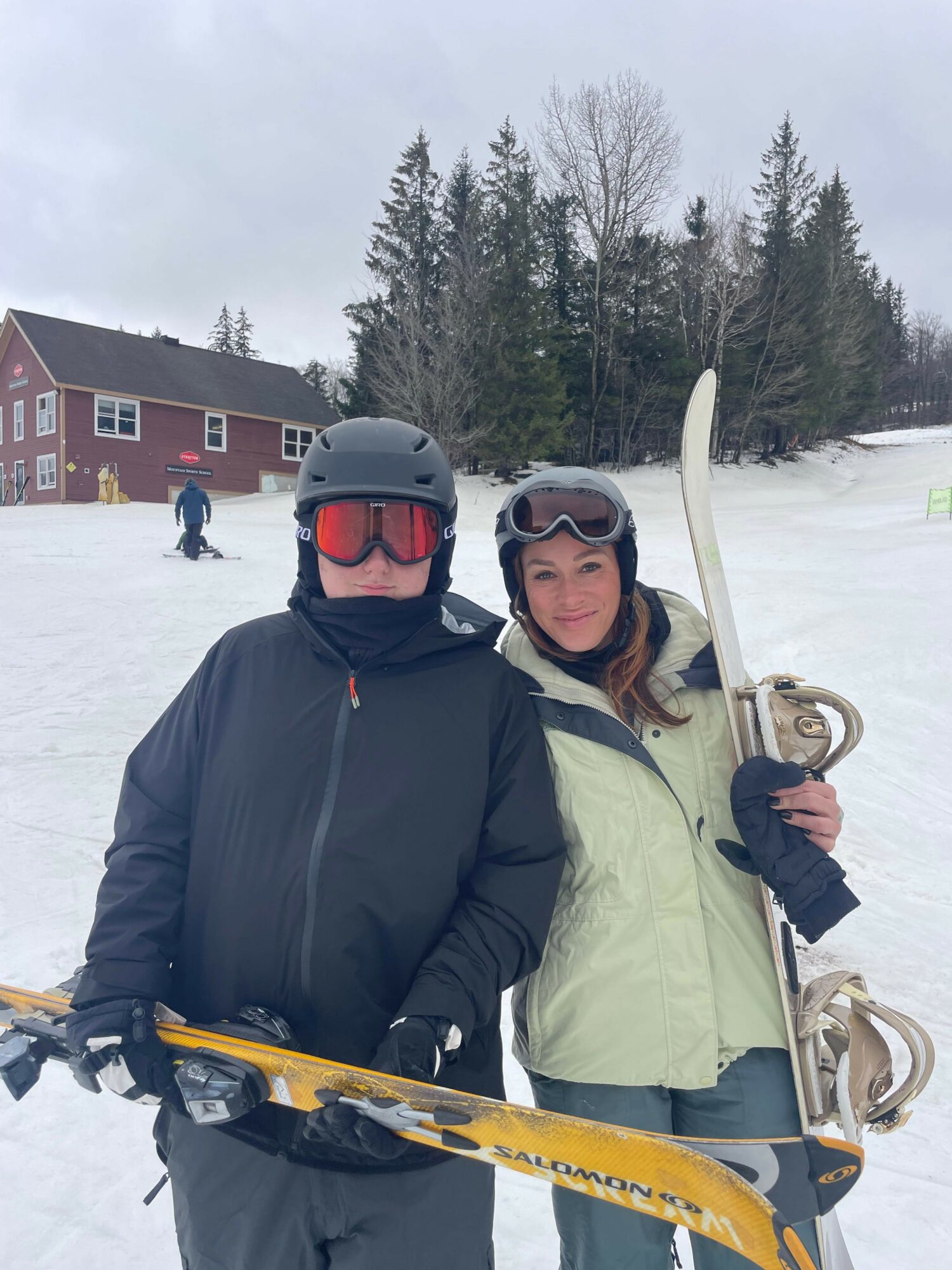 Two people in winter clothing and ski gear standing on snow, with trees and a building in the background.