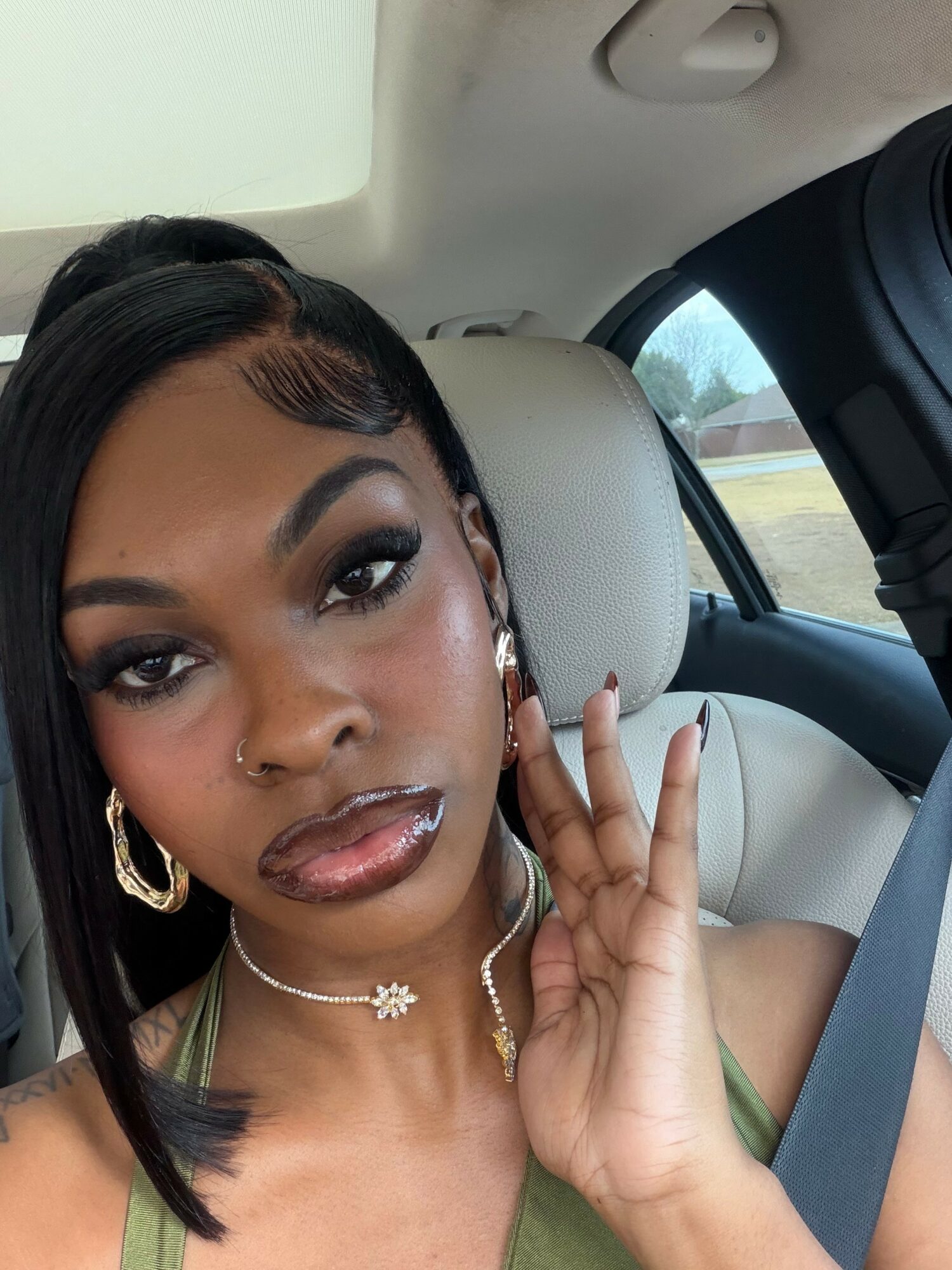 Woman with dark hair, makeup, jewelry, sitting in car, holding hand near face, looking at camera.