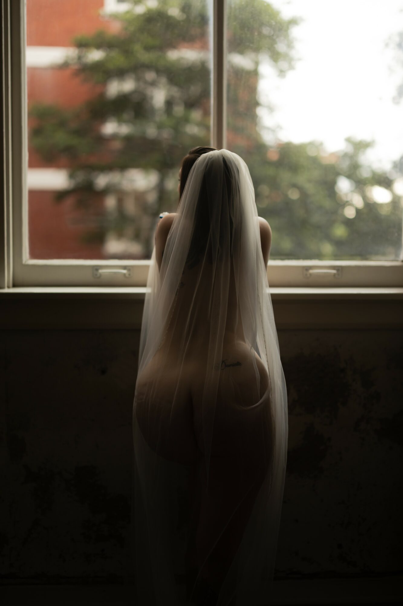 A person with long hair sitting on a windowsill, looking outside at trees and buildings, with natural light illuminating the scene.