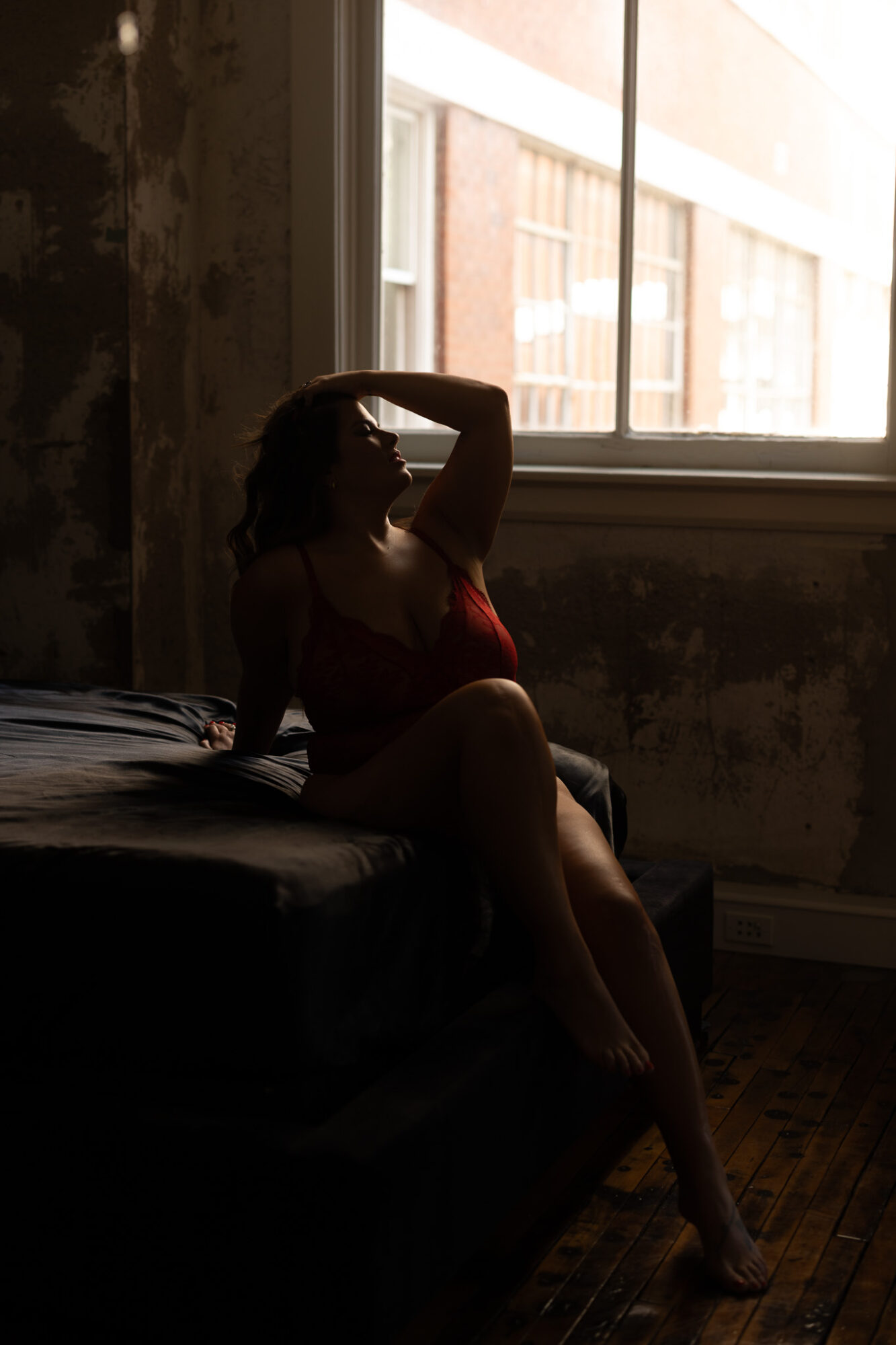 Silhouette of a woman sitting on a bed near a window, with sunlight illuminating the background, dark interior, relaxed pose.