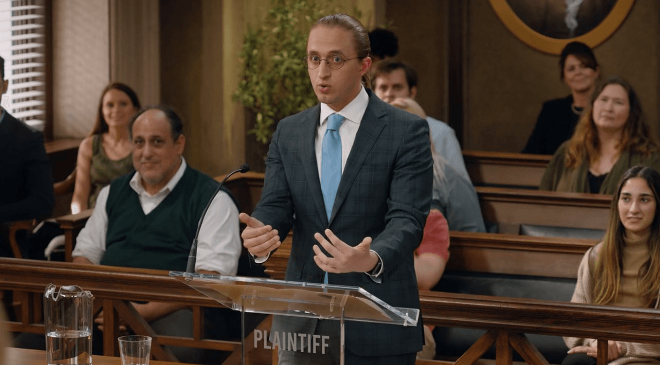 Man in suit speaking in courtroom with seated audience behind him.
