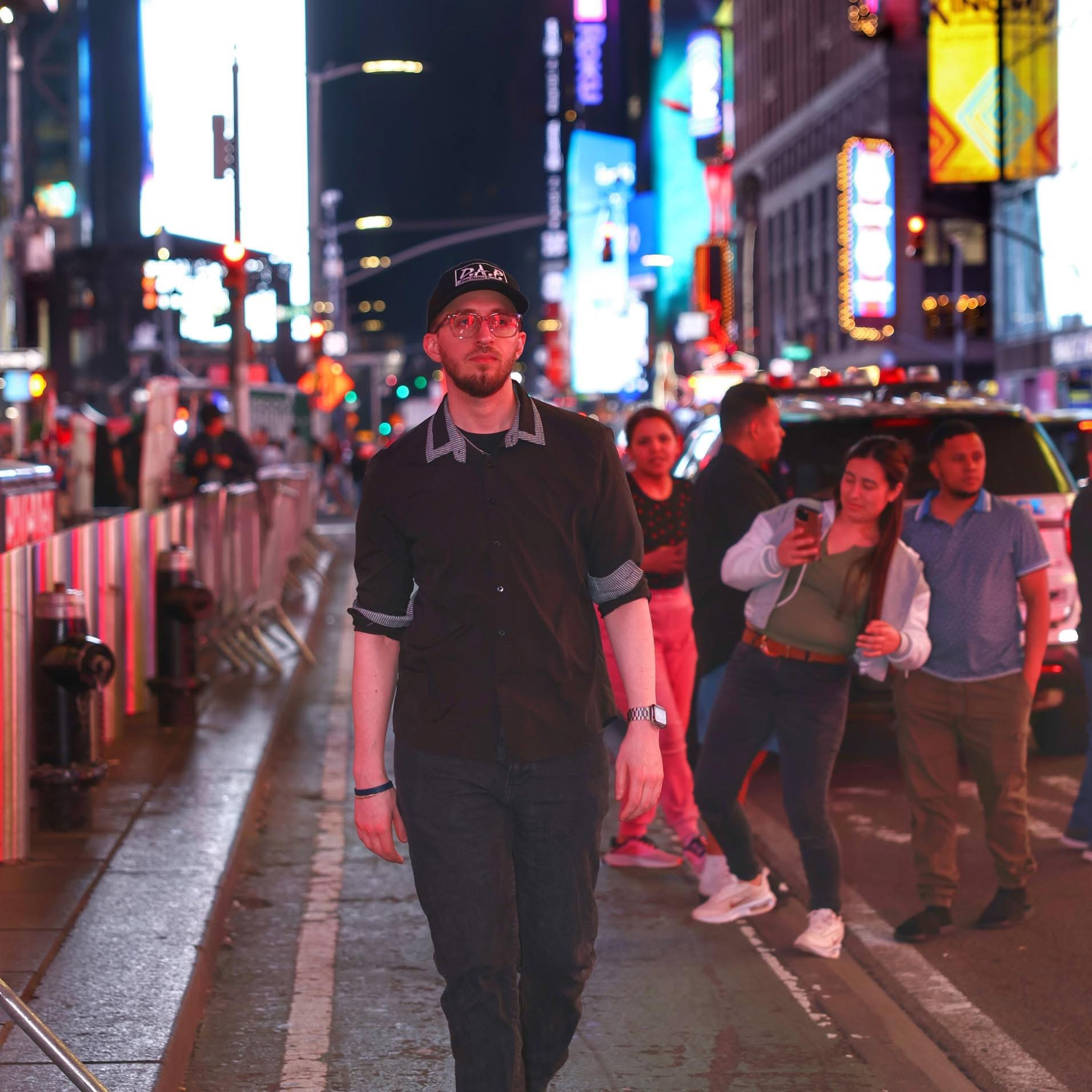 Man walking on city street at night with bright lights and pedestrians around, wearing glasses, a cap, and dark clothing.