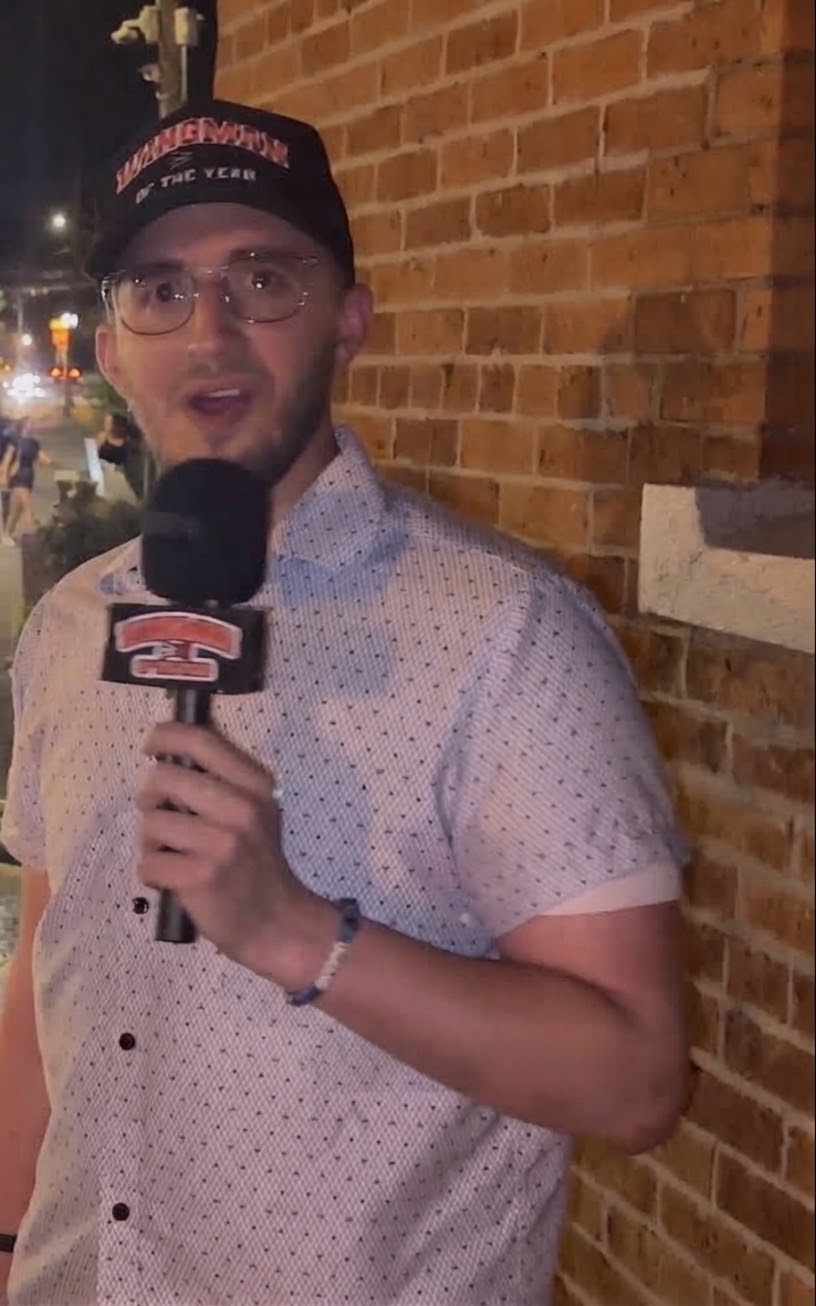 Man holding microphone in front of brick wall, wearing glasses and a cap, speaking outdoors at night.